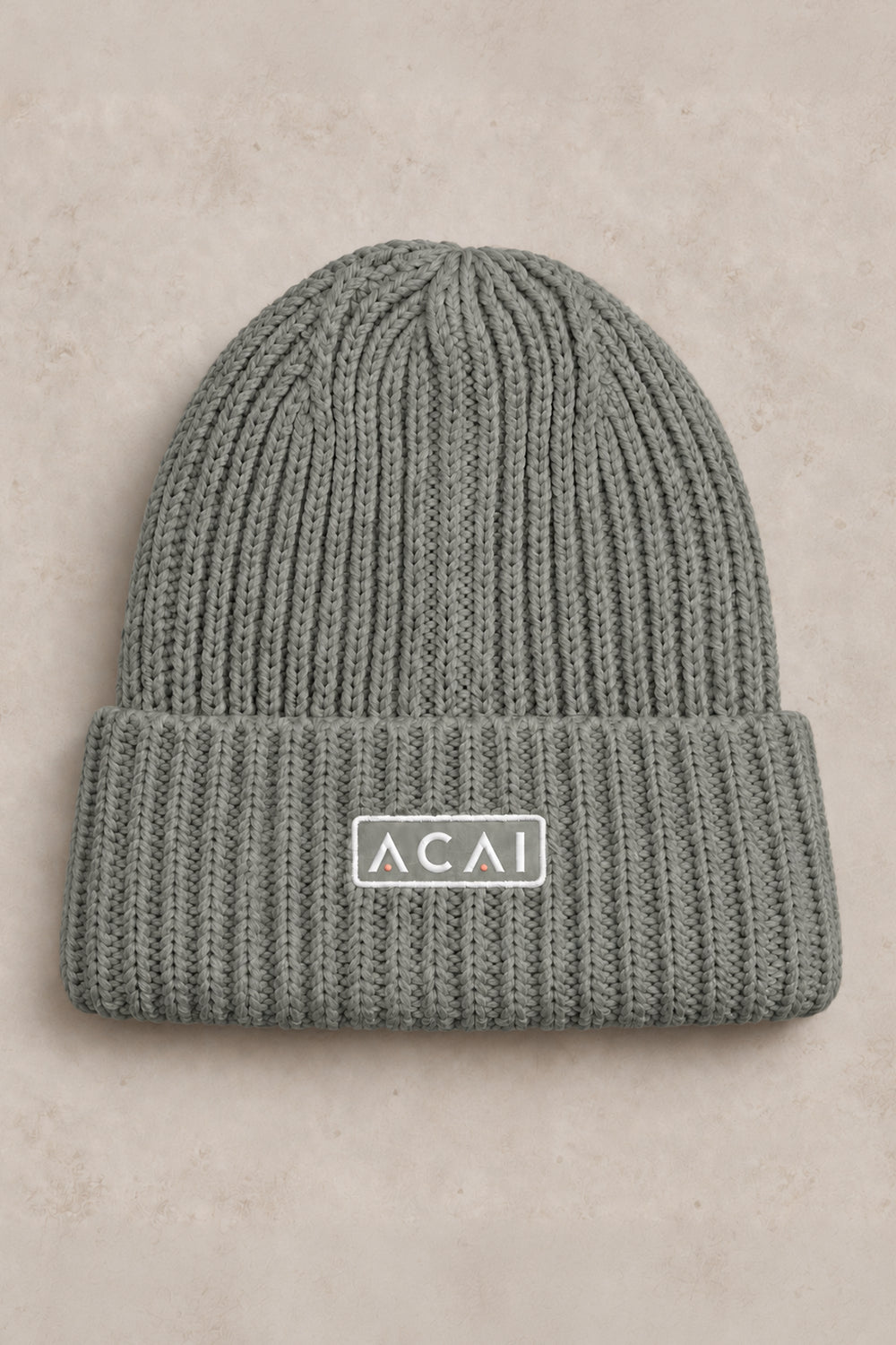 A ribbed knit beanie hat in a muted sage green color, featuring a folded cuff with a white rectangular patch that reads 'ACAI' in white letters. The hat is photographed flat against a textured, light beige background.
