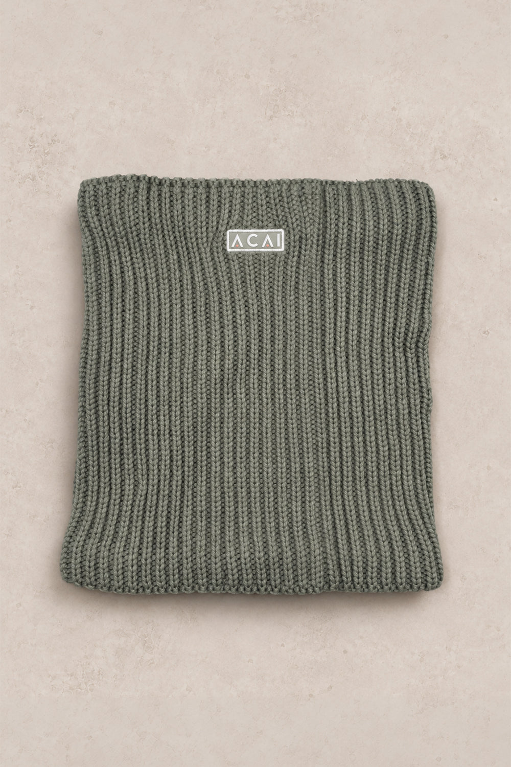 A folded ribbed knit beanie in a muted sage green color, featuring a small white rectangular label with the brand name 'ACAI' on the front, set against a light beige textured background.