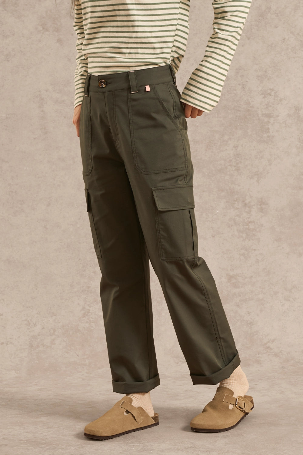 A person wearing olive green cargo pants with a cream and green striped long-sleeve shirt and tan clogs, standing against a textured beige studio backdrop.