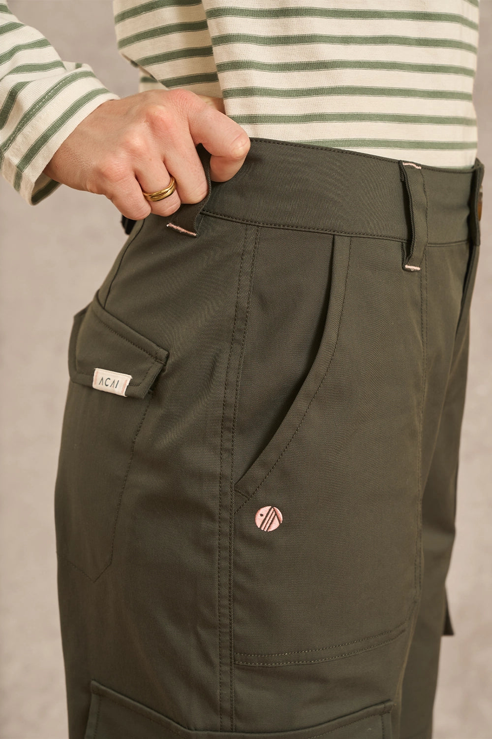 Close-up of a person wearing olive green cargo-style pants, with a striped long-sleeve shirt partially visible. A small circular logo patch and a small fabric brand tab are visible on the pants.