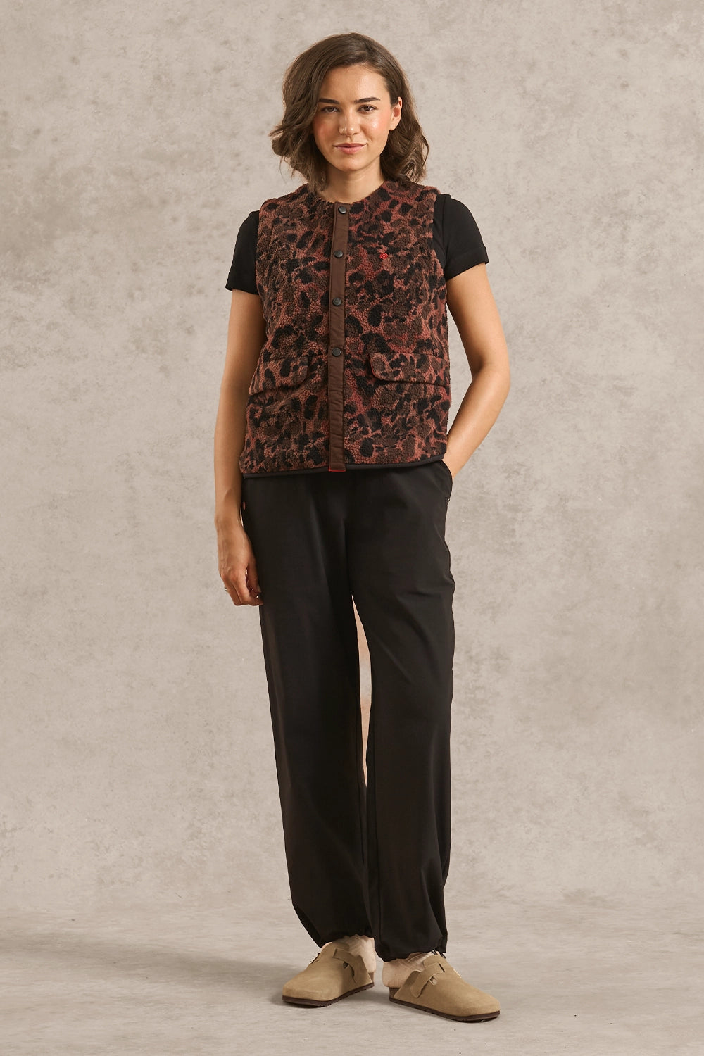 A woman stands against a textured light gray studio backdrop wearing a sleeveless cropped vest with a brown and black animal-style pattern over a black short-sleeve top, black wide-leg pants, and tan shoes.