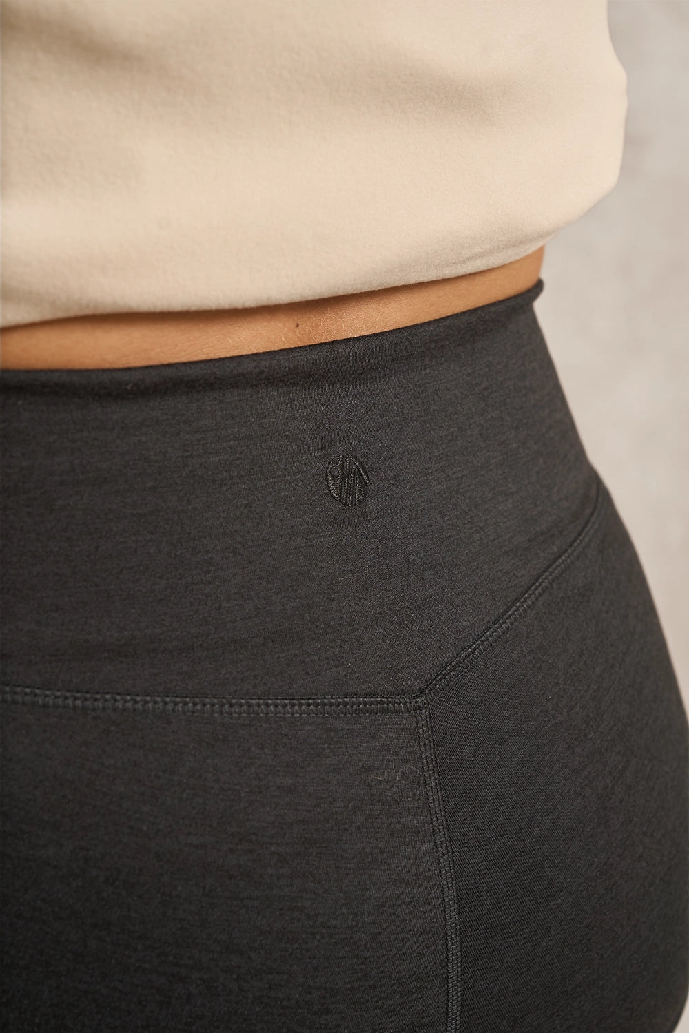 Close-up of a person wearing dark charcoal gray high-waisted leggings with a small tonal logo at the center back waist, paired with a light beige top against a neutral gray background.
