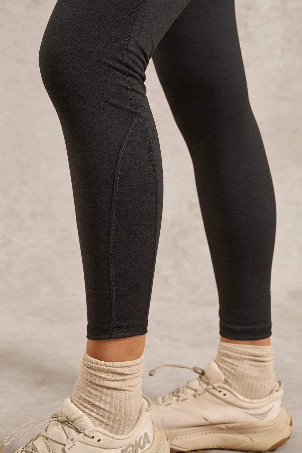 Close-up of a person's legs wearing black athletic leggings, beige ribbed ankle socks, and beige sneakers against a neutral textured studio background.