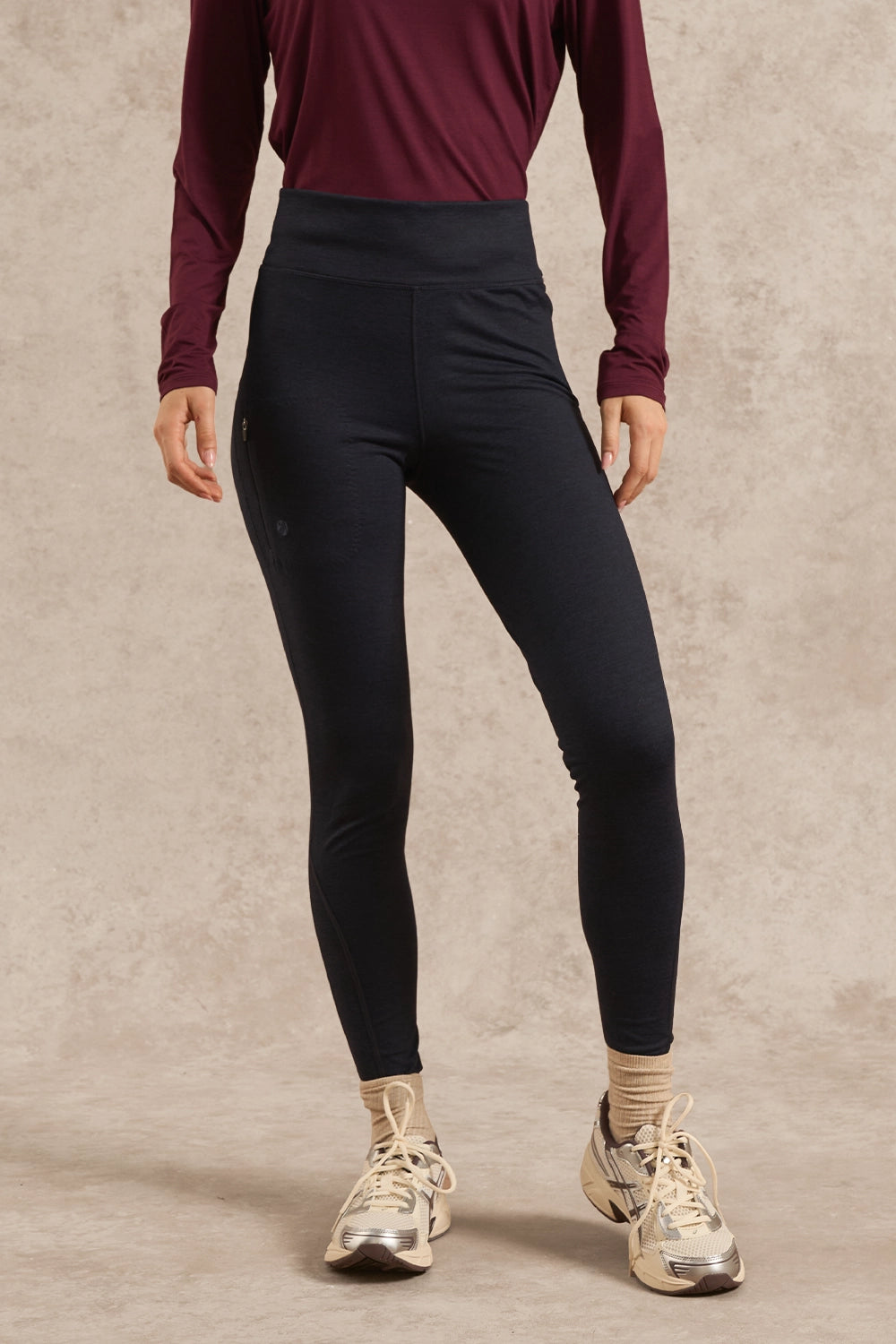 A model wearing high-waisted black athletic leggings, a burgundy long-sleeve top, beige socks, and metallic sneakers, standing against a textured beige studio backdrop.