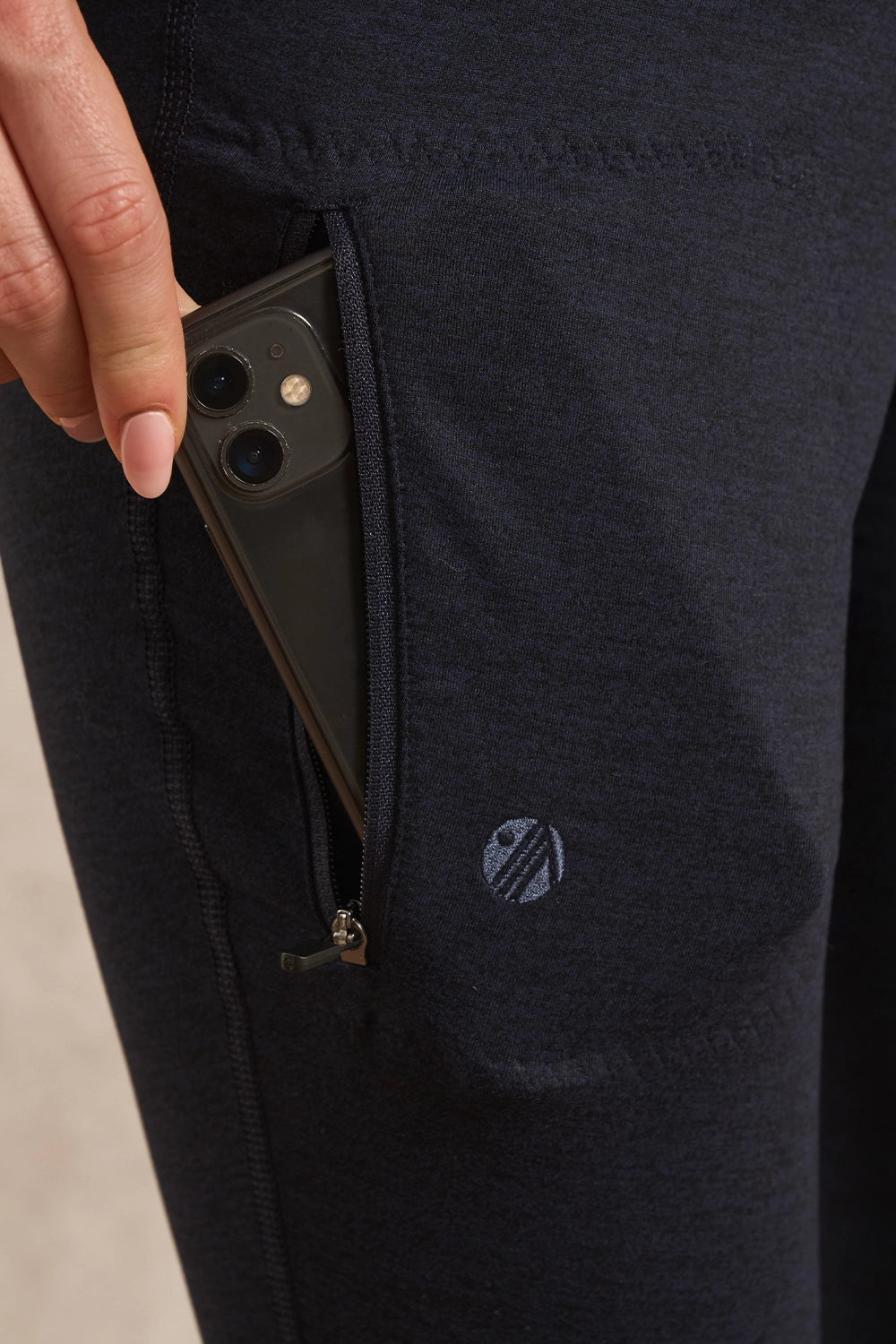 Close-up of a person's hand pulling a smartphone out of a side pocket of dark navy heathered athletic pants. A small circular embroidered logo is visible on the leg.