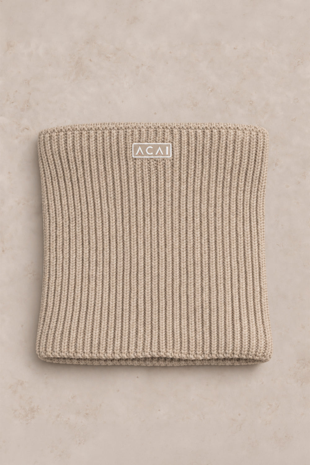 A folded ribbed knit beanie in a warm beige/tan color, featuring a small white rectangular label with the brand name 'ACAI' in black text, set against a matching textured beige background.