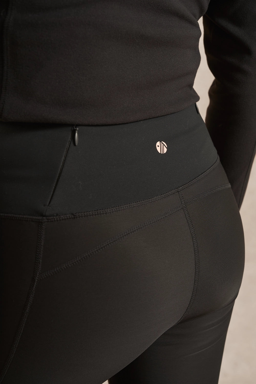 Close-up of a person wearing black athletic leggings with a small metallic logo near the waistband, paired with a black long-sleeve top.