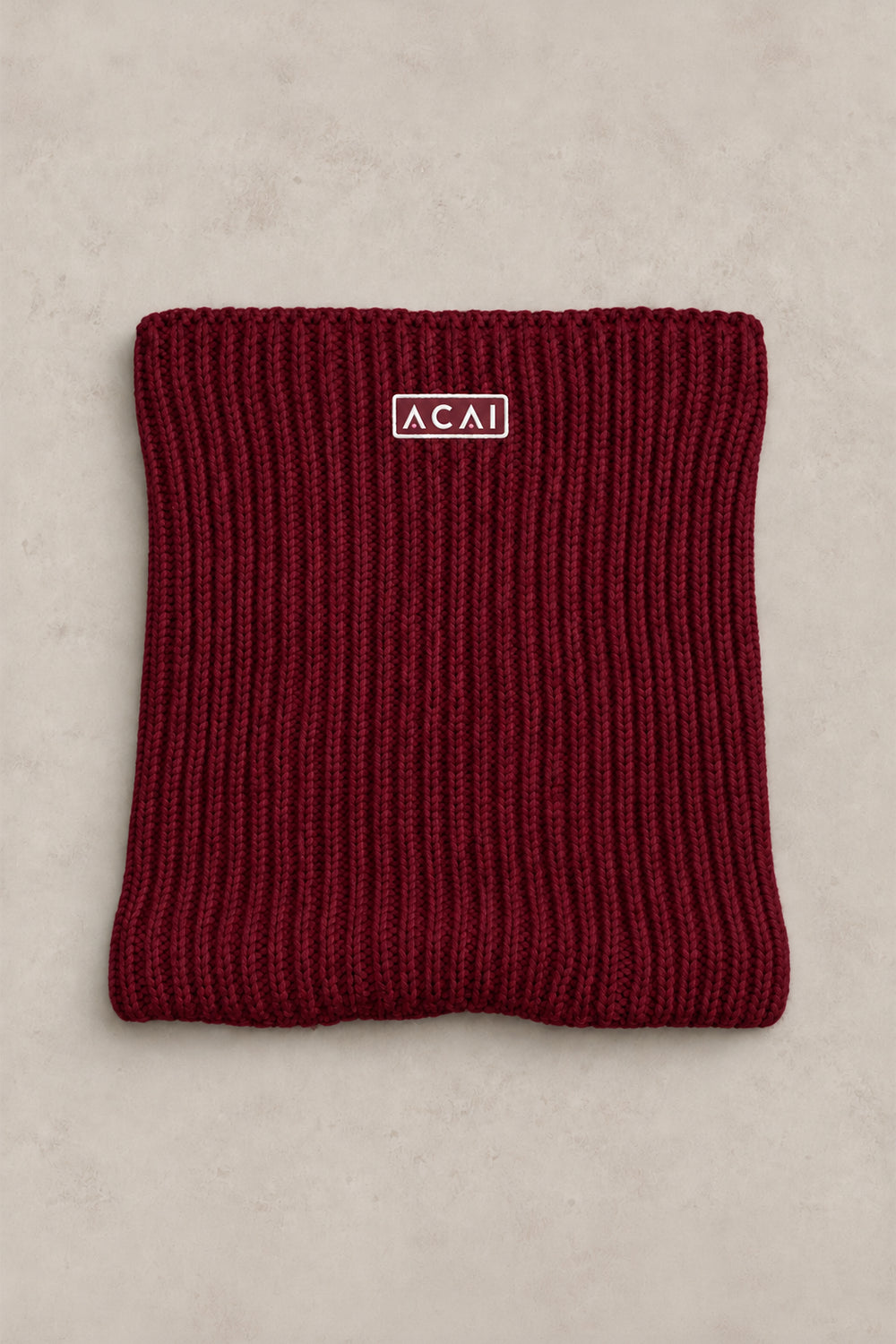 A folded ribbed knit beanie in deep burgundy red, featuring a small white rectangular logo patch with the brand name 'ACAI' on the front, set against a neutral light beige textured background.
