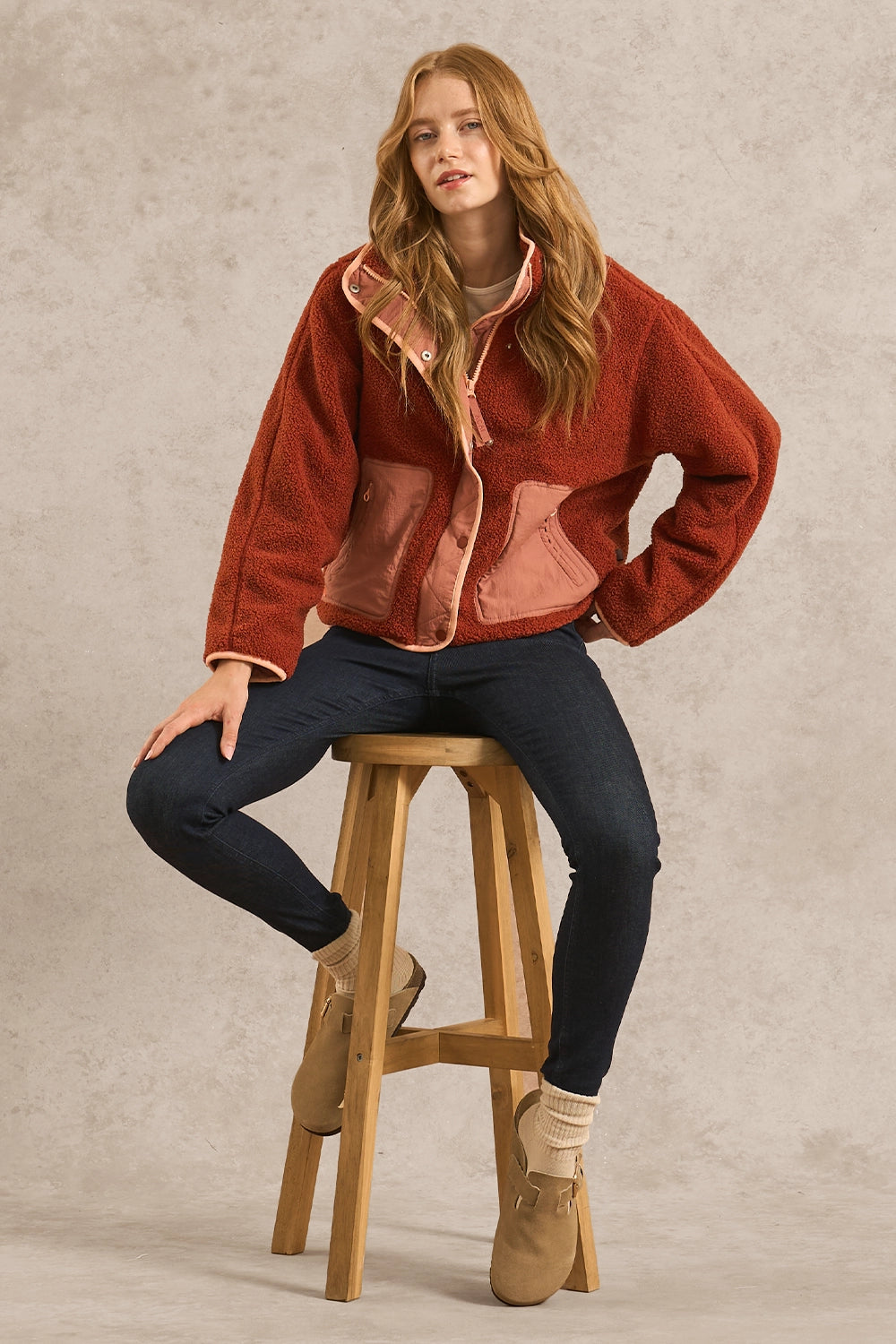 A woman sits on a wooden stool wearing a rust-orange sherpa fleece jacket with dusty pink lining, dark blue jeans, and tan boots against a textured beige studio backdrop.
