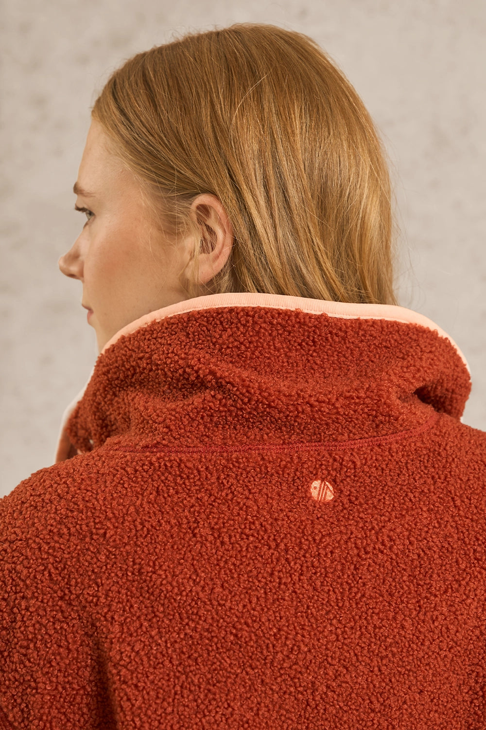 A close-up back view of a woman wearing a textured terracotta red fleece jacket with a light pink inner collar, set against a neutral gray background.
