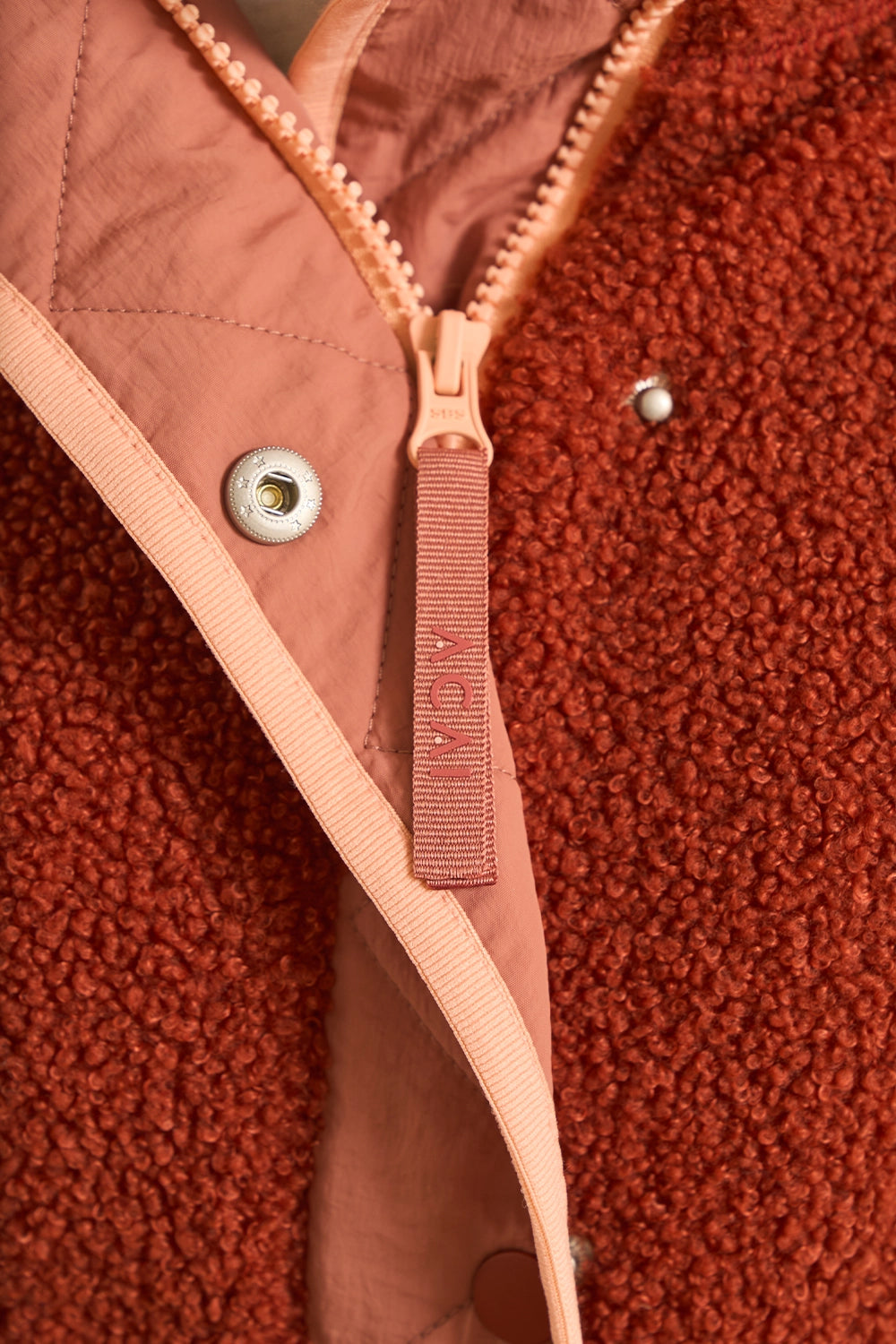 Close-up of a fleece jacket with a terracotta rust-brown sherpa exterior and a dusty pink inner lining and zipper pull tab. The zipper pull tab is embroidered with the brand name 'MCM'.