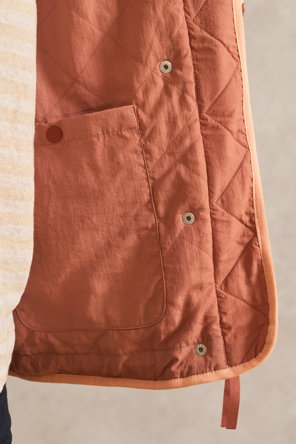 Close-up of a quilted rust-orange vest with diamond stitching, a front pocket, and metal snap buttons, shown against a neutral beige background.