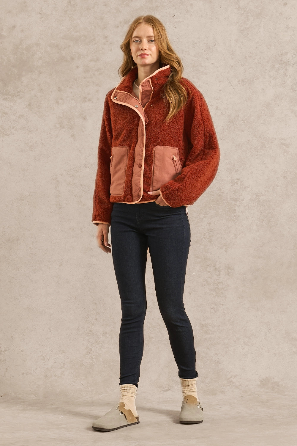 A woman stands against a textured beige studio backdrop wearing a rust-orange sherpa-lined jacket with dusty pink front pockets, dark navy skinny jeans, cream socks, and light gray slip-on shoes.