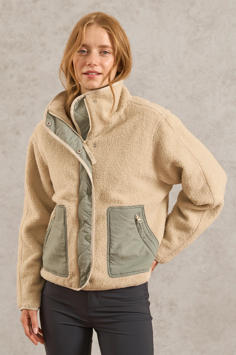 A woman wearing a beige sherpa fleece jacket with sage green contrast panels and dark gray pants, standing against a textured gray wall.