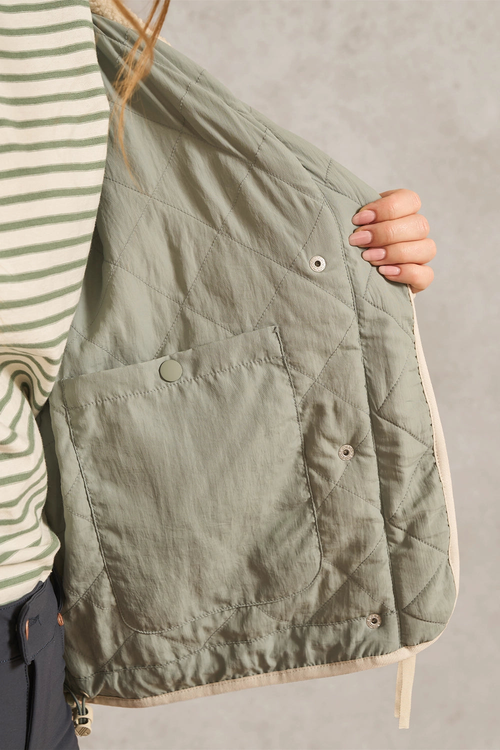 A close-up of a person holding up an olive green quilted jacket or vest, showing the interior pocket and buttoned closure. The person is wearing a green and white striped shirt, and the background is a neutral gray wall.