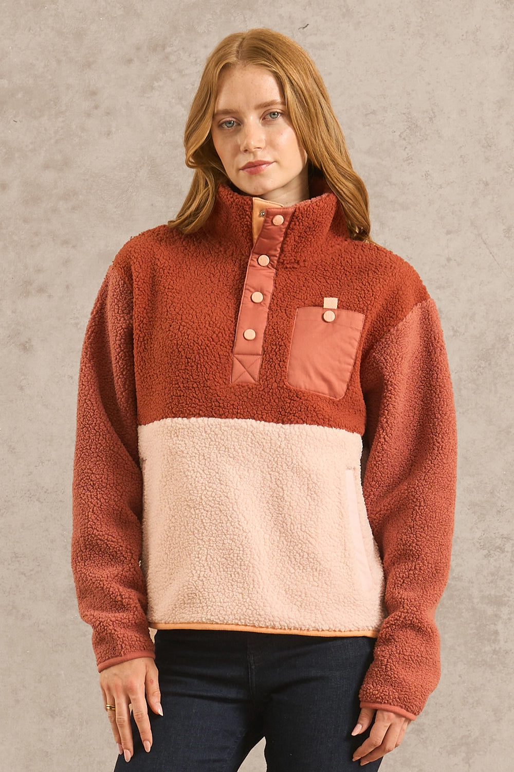 A woman modeling a colorblock fleece pullover sweater with a rust orange top half, a pale pink lower half, and matching chest pocket details, paired with dark skinny jeans and dark brown boots against a textured gray studio backdrop.