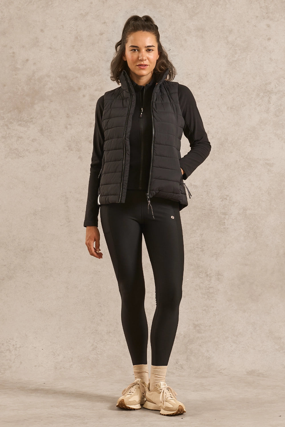 A woman modeling a black puffer vest over a black long-sleeve top and black leggings, standing against a textured beige studio backdrop.