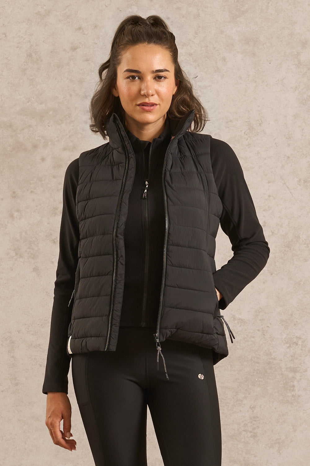 A woman modeling a black quilted zip-up puffer vest over a black long-sleeve top and black leggings, standing against a textured light gray studio background.