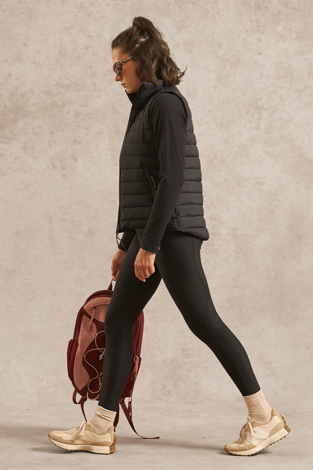 A woman in a black puffer vest, black leggings, and beige sneakers walks while carrying a burgundy backpack against a textured beige studio backdrop.