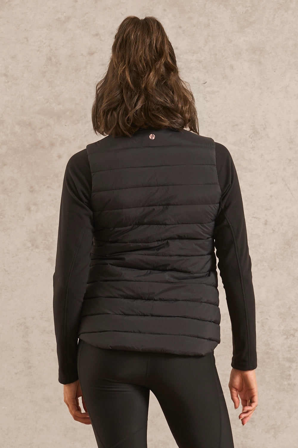 A woman viewed from behind wearing a black quilted puffer vest over a black long-sleeve top and black leggings, standing against a textured light gray wall.