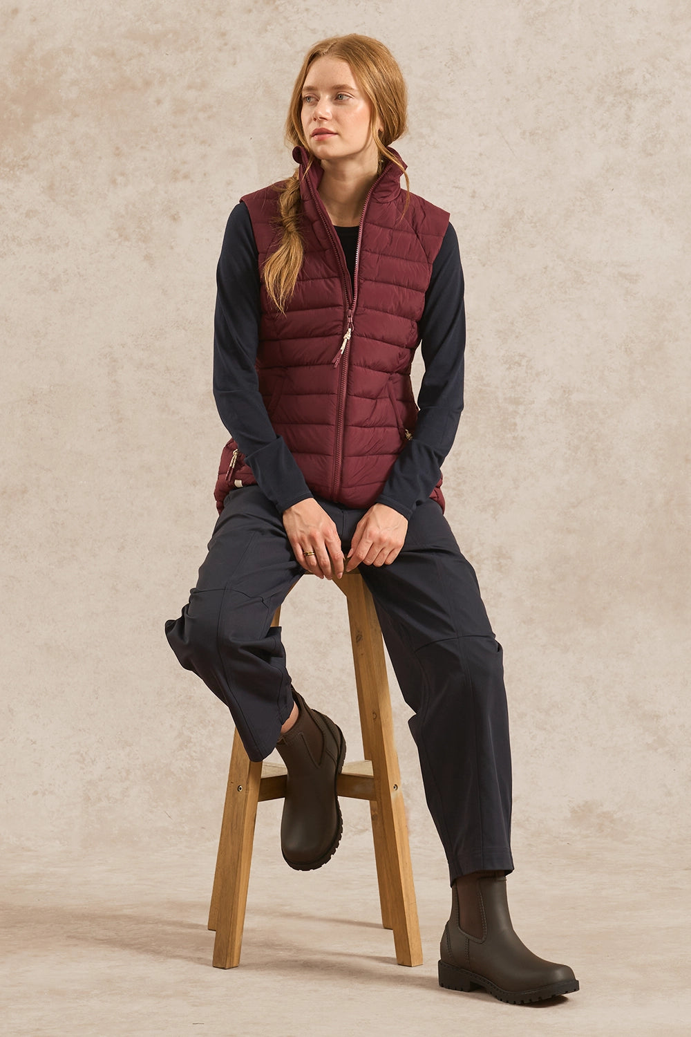 A woman sits on a wooden stool wearing a burgundy quilted puffer vest over a dark navy long-sleeve top, navy pants, and dark boots against a textured beige studio backdrop.