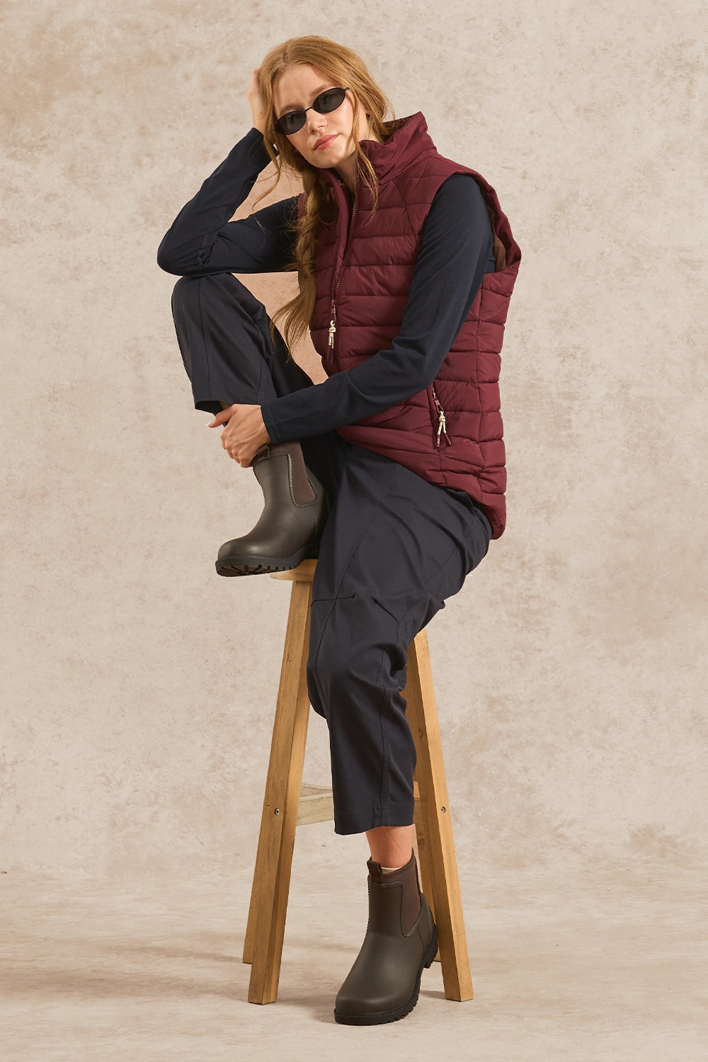 A woman wearing a burgundy quilted puffer vest over a navy long-sleeve top and navy pants, sitting on a wooden stool against a textured beige studio backdrop.