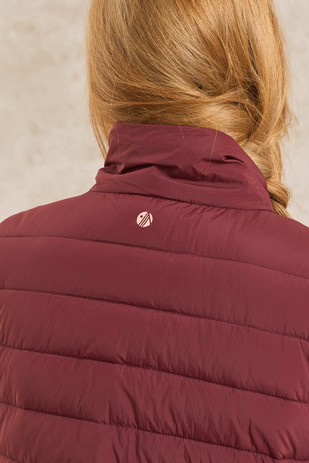 Close-up back view of a person wearing a quilted puffer jacket in burgundy with a small circular logo on the upper back.
