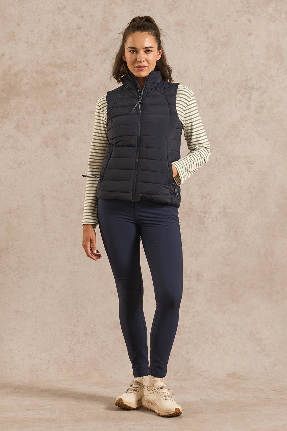 A woman modeling a dark navy sleeveless puffer vest over a cream and navy striped long-sleeve shirt, paired with navy leggings and beige sneakers, against a textured beige studio backdrop.