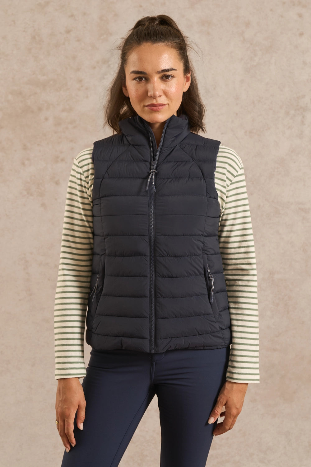 A woman modeling a dark navy quilted puffer vest over a cream and navy striped long-sleeve shirt, paired with navy pants, against a textured beige studio backdrop.