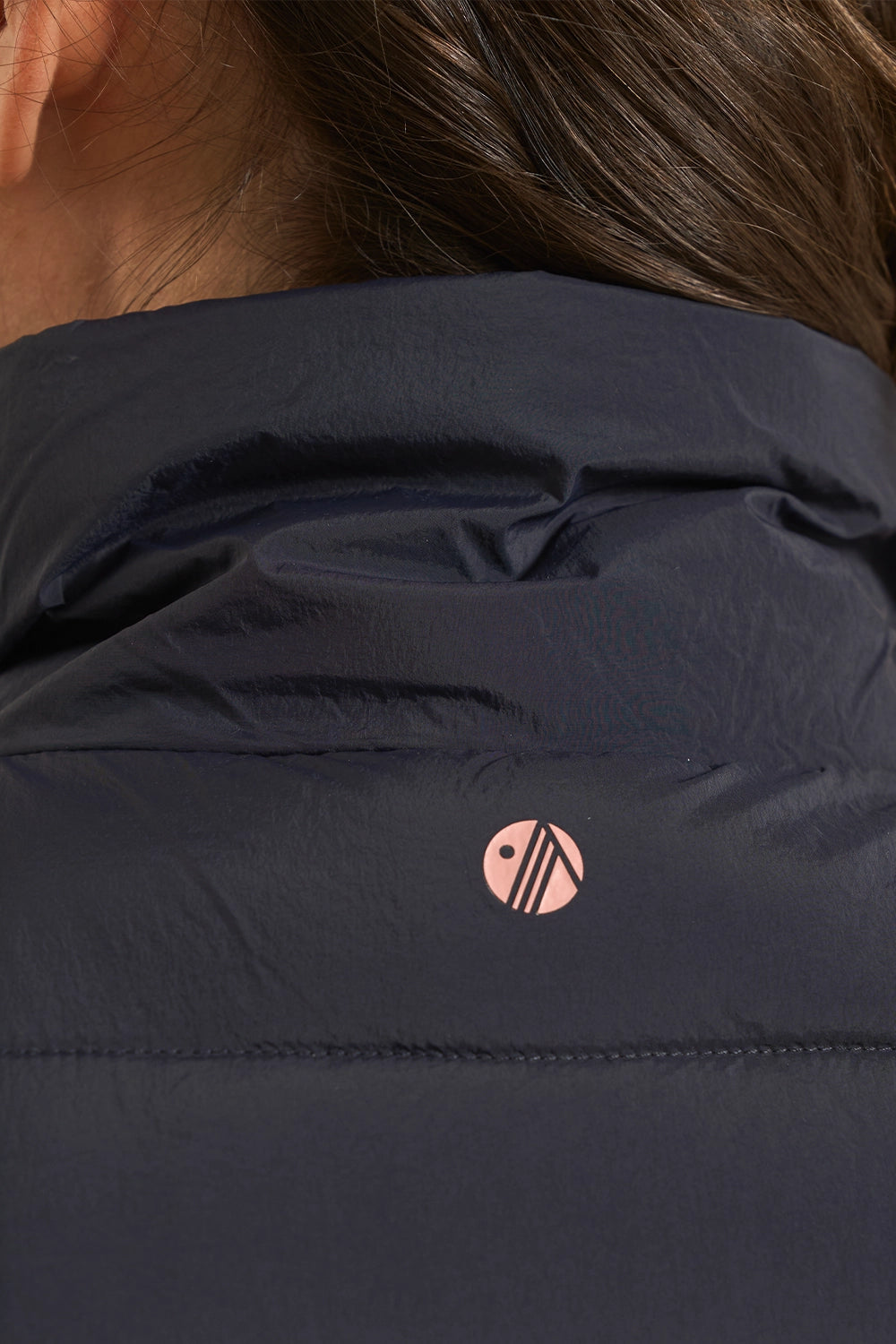 Close-up of a person wearing a dark navy puffer jacket, showing the back of the neck, hair, and a small pink circular logo on the upper back.