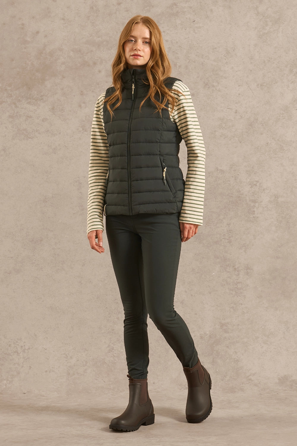 A woman modeling a dark gray zip-up puffer vest over a cream and gray striped long-sleeve shirt, paired with dark gray skinny pants and dark brown boots, against a textured beige studio backdrop.