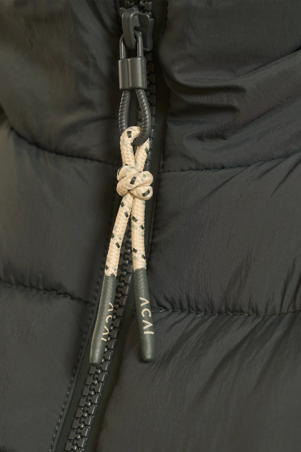 Close-up of a black puffer jacket zipper with a cream and black speckled rope pull tied in a knot, featuring the brand name ACAI printed on the metal tips.