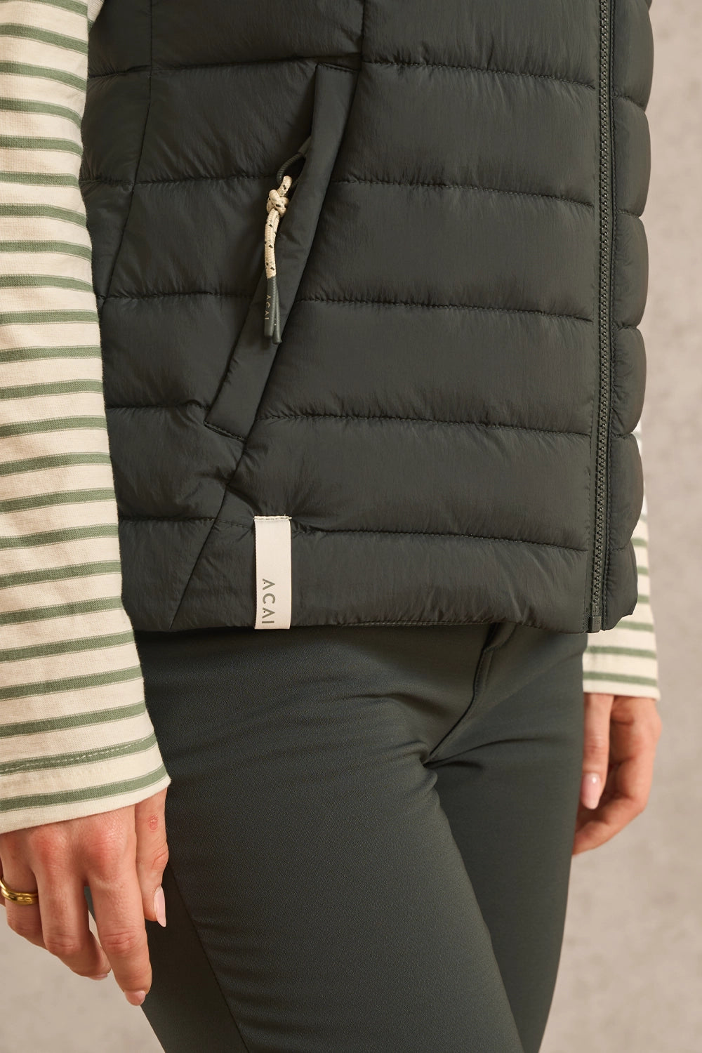 Close-up of a person wearing a black puffer vest over a green and white striped long-sleeve top and dark leggings. A small white fabric tag on the vest reads AEAU.