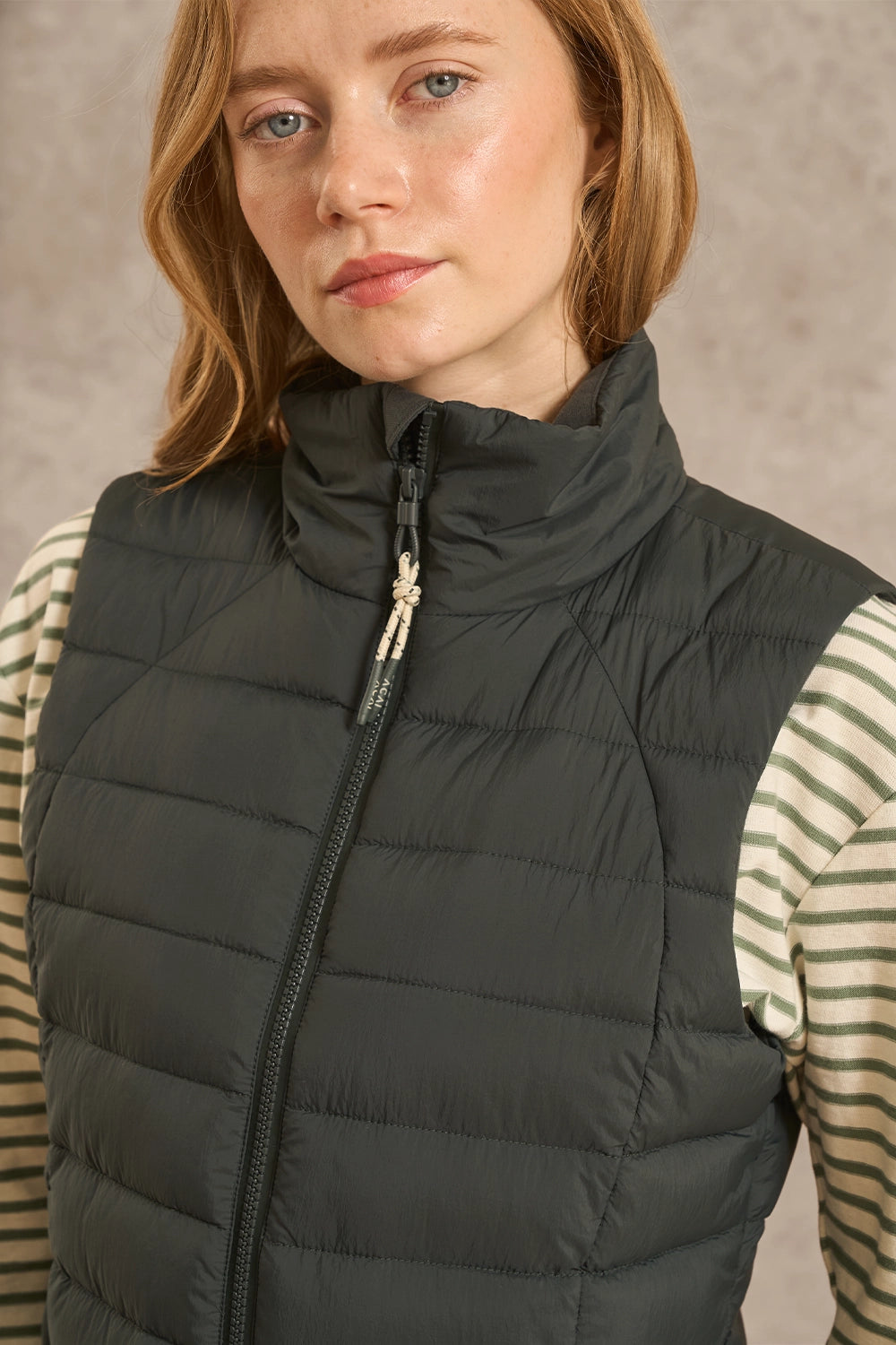 A woman wearing a dark gray quilted puffer vest over a green and white striped long-sleeve shirt, photographed against a neutral textured background.