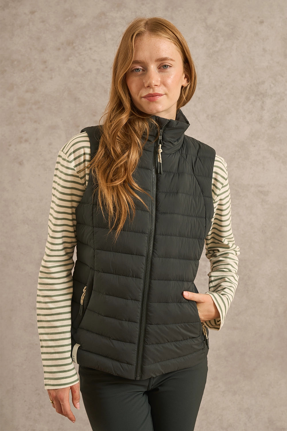 A woman wearing a dark gray quilted puffer vest over a cream and gray striped long-sleeve shirt, standing against a textured gray studio background.