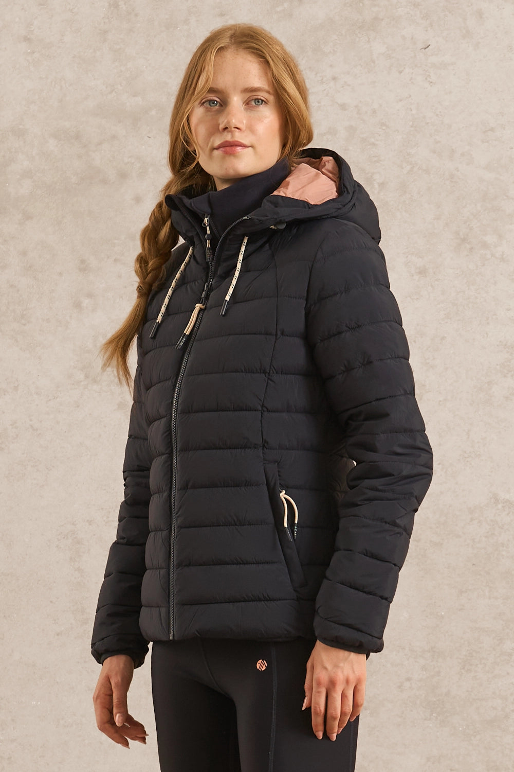 A woman wearing a black hooded puffer jacket with light beige drawstrings, standing against a textured beige wall.