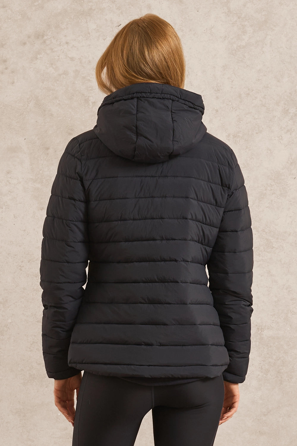 A woman viewed from the back wearing a black quilted puffer jacket with a hood, set against a textured light beige wall.