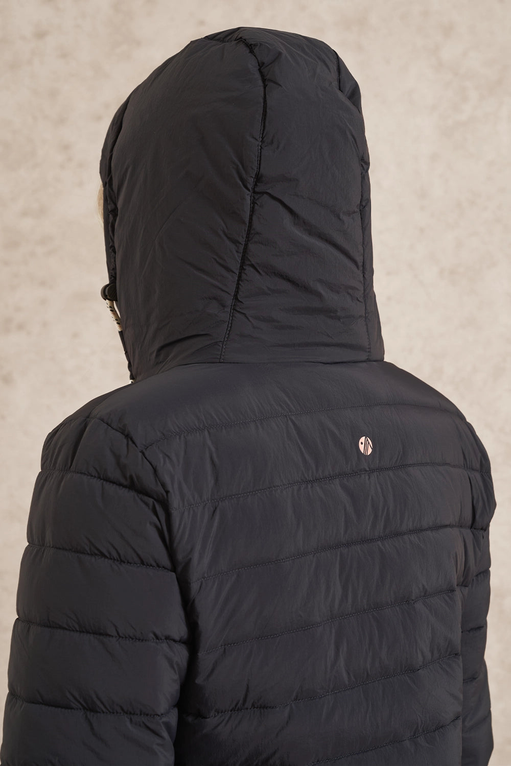 Close-up back view of a black hooded puffer jacket with horizontal quilting and a small circular logo near the upper right shoulder, shown against a textured beige background.