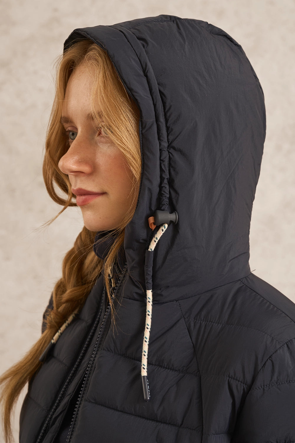A close-up side profile of a woman wearing a dark navy hooded puffer jacket against a textured light beige background.