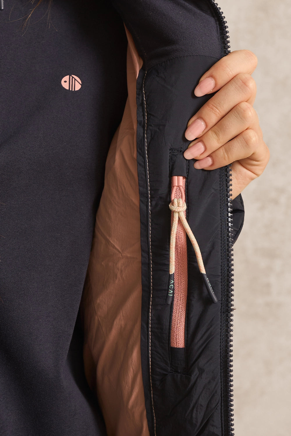 Close-up of a person wearing a dark navy jacket with a brown quilted lining, featuring a zippered pocket with a pink pull tab and a beige drawstring, against a neutral textured background.