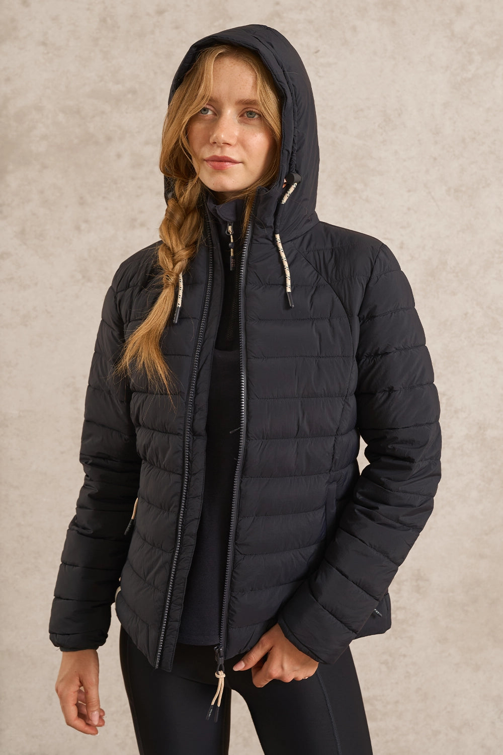 A woman wearing a black hooded puffer jacket with a front zipper and light-colored drawstrings, standing against a textured beige studio background.