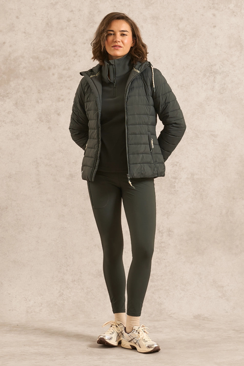 A woman modeling a dark olive green quilted puffer jacket over a matching dark green turtleneck and leggings, paired with beige sneakers, against a textured beige studio backdrop.