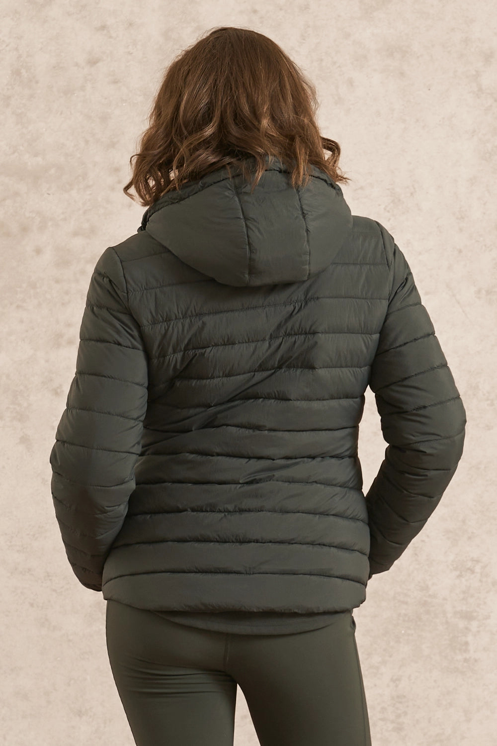 A woman viewed from behind wearing a dark olive green hooded puffer jacket and matching dark leggings against a textured beige wall.