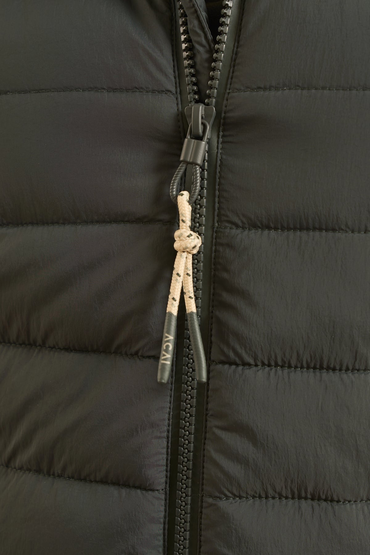 Close-up of a black puffer jacket with horizontal quilting, a black zipper, and a cream-colored drawstring zipper pull featuring a knot and the brand name 'ACAI' printed on it.