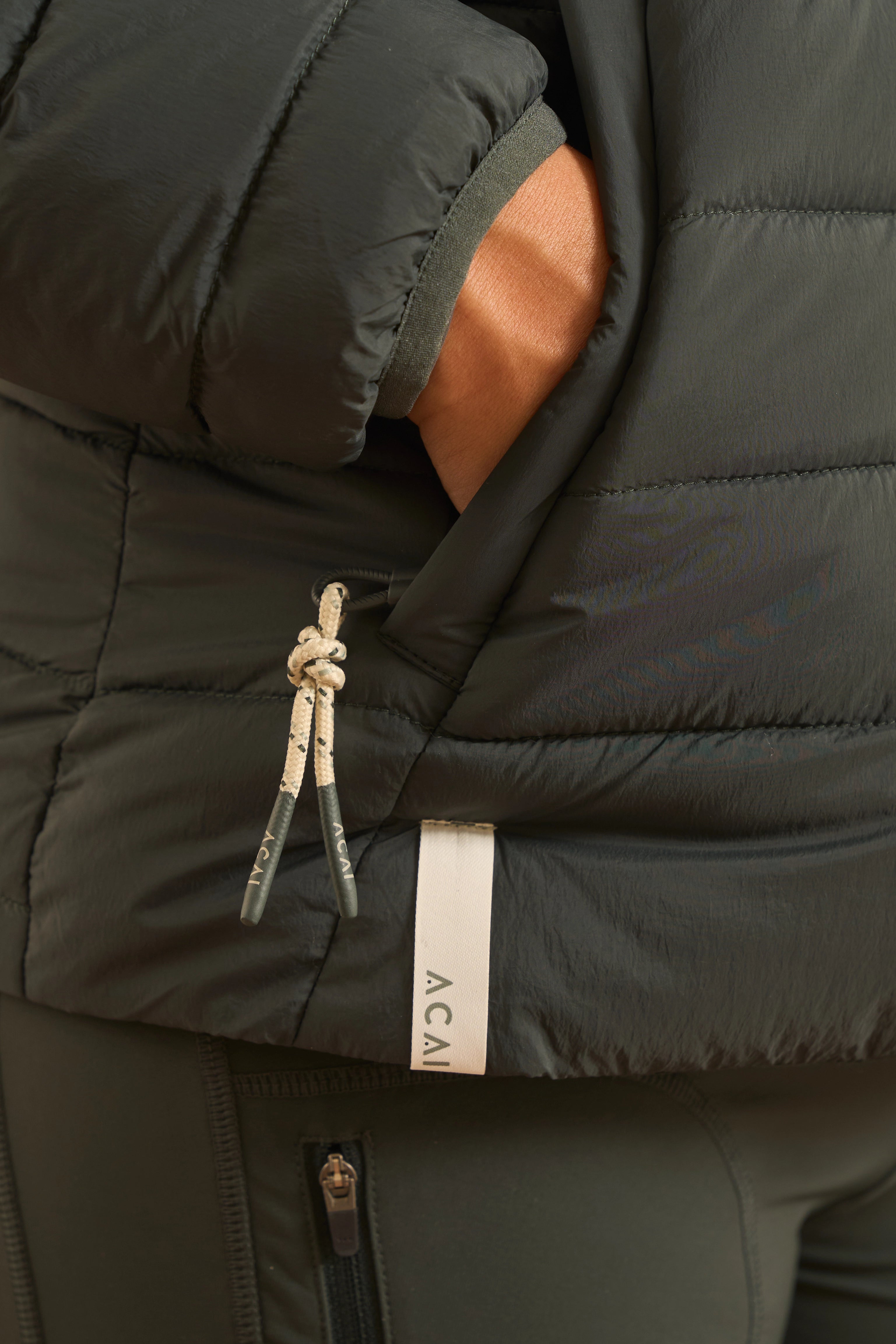 Close-up of a dark olive green puffer jacket with a white and gray patterned drawstring and a white vertical brand tag reading ACAI.