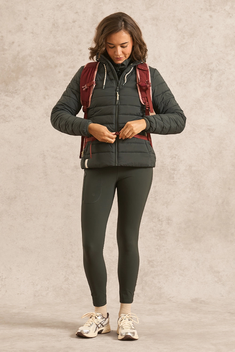 A woman wearing a dark gray puffer vest over a burgundy backpack, olive green leggings, and white sneakers, standing against a textured beige studio backdrop.