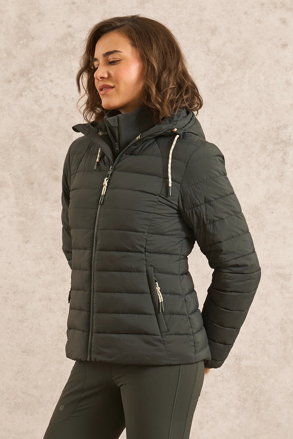 A woman wearing a dark olive green hooded puffer jacket with silver zippers, standing against a textured light beige wall.