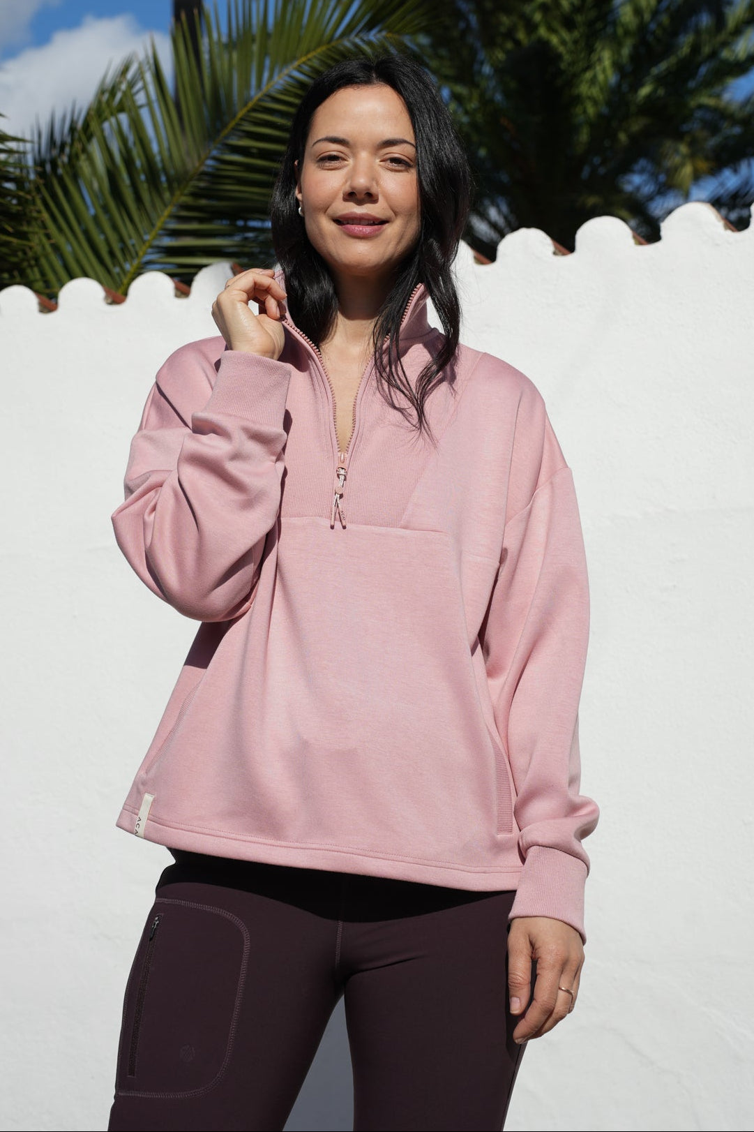 Woman wearing a pink long-sleeve top standing in front of a white fence with palm trees in the background.