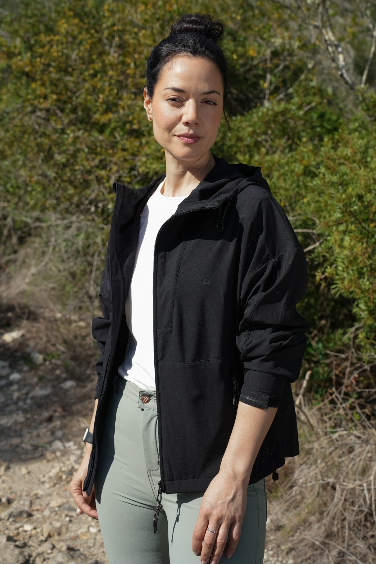 Woman wearing a black jacket and light gray pants standing outdoors with greenery in the background