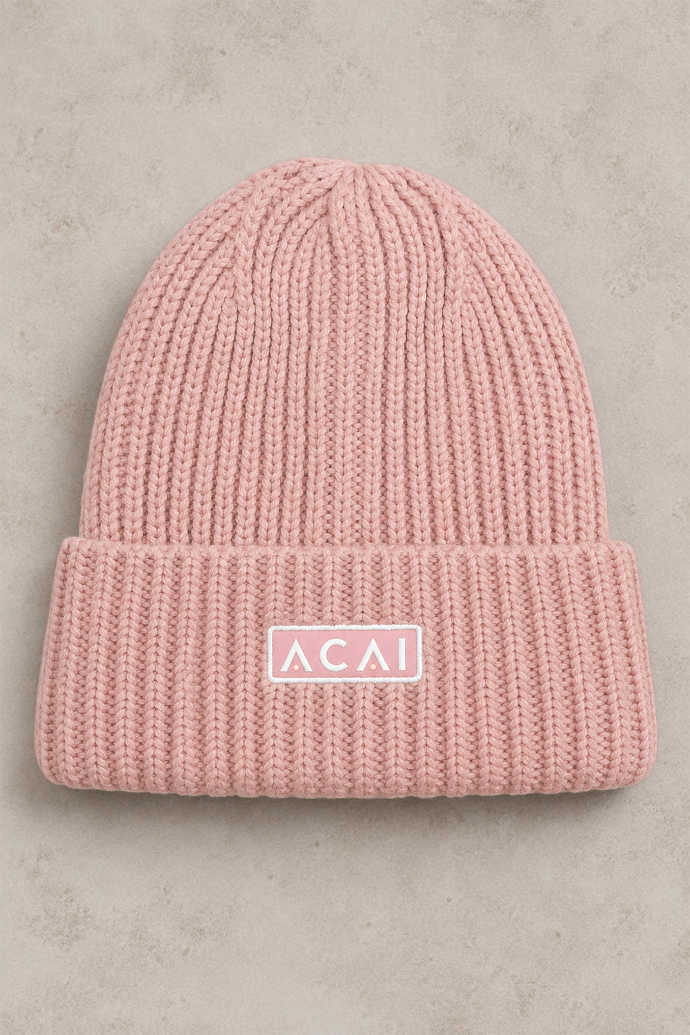 A light pink ribbed knit beanie hat with a folded cuff, featuring a white rectangular patch with the text 'ACAI' in white. The hat is photographed flat against a textured, light gray background.